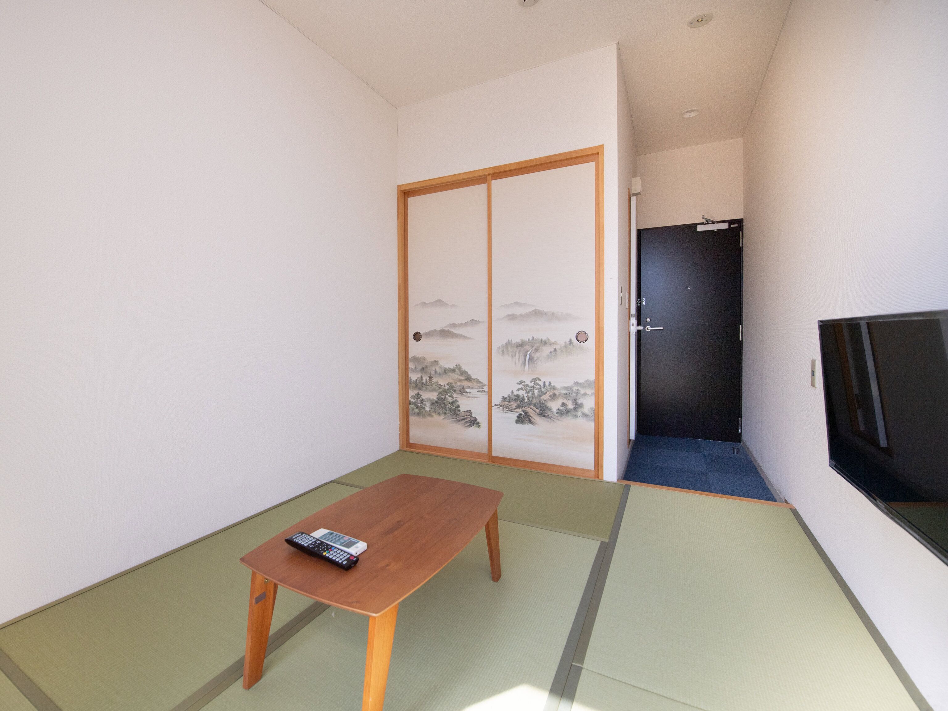 A small room with tatami flooring, a wooden table with a remote control, a sliding shoji screen door, and a dark door leading to another room.