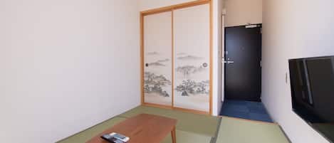 A small room with tatami flooring, a wooden table with a remote control, a sliding shoji screen door, and a dark door leading to another room.
