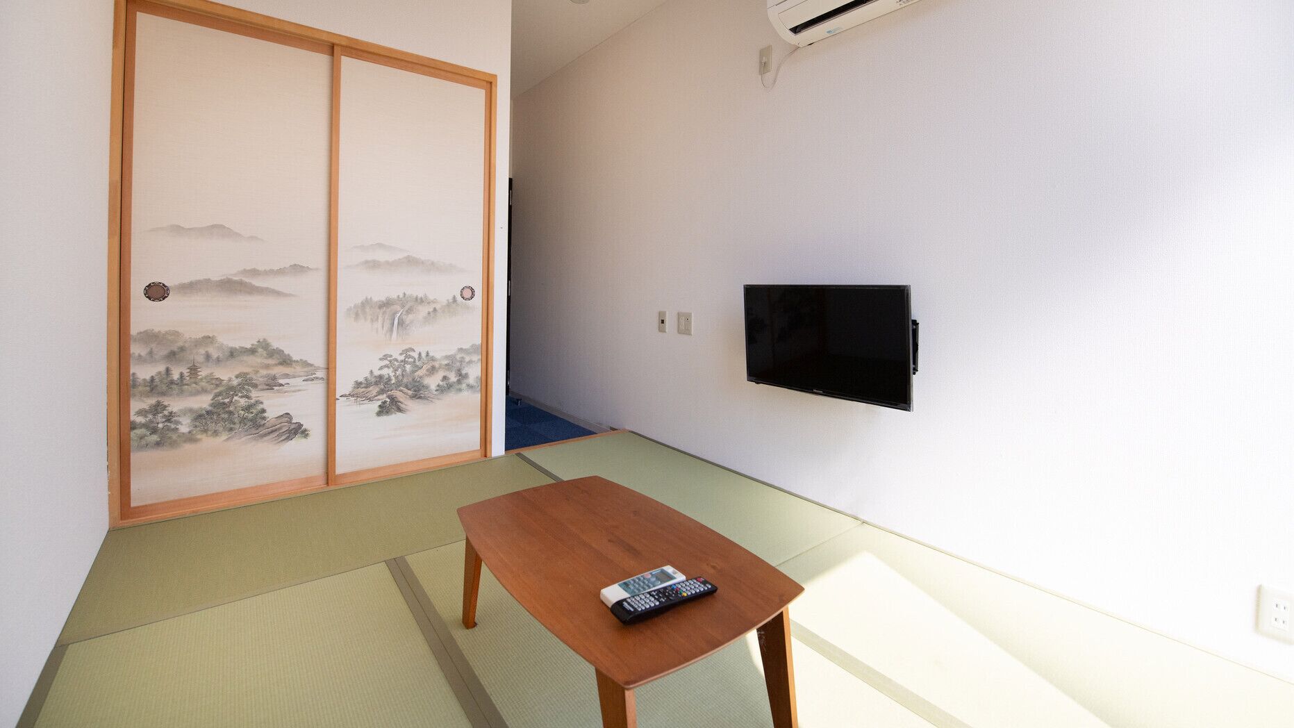 Una habitación con suelo de tatami, un panel corredizo shoji, un televisor montado en la pared y una pequeña mesa de madera con un control remoto.