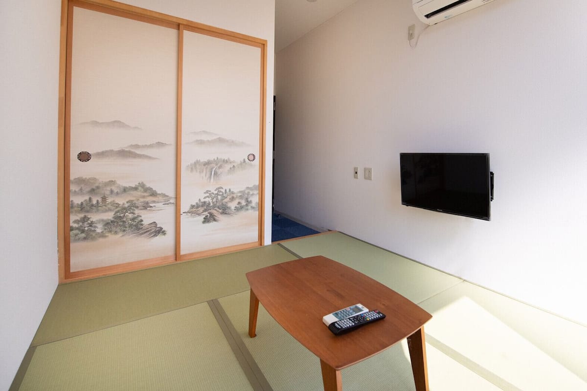 Una habitación con suelo de tatami, un panel corredizo shoji, un televisor montado en la pared y una pequeña mesa de madera con un control remoto.