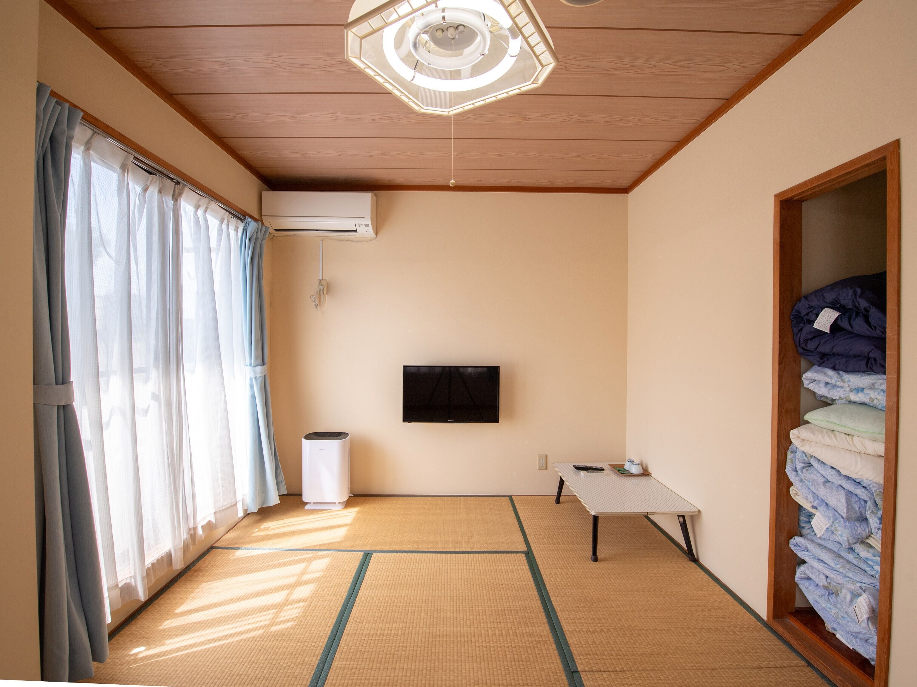 A room with tatami mat flooring, a ceiling fan, an air conditioning unit, a television mounted on the wall, a small table with a remote control, and a wardrobe with folded clothes.
