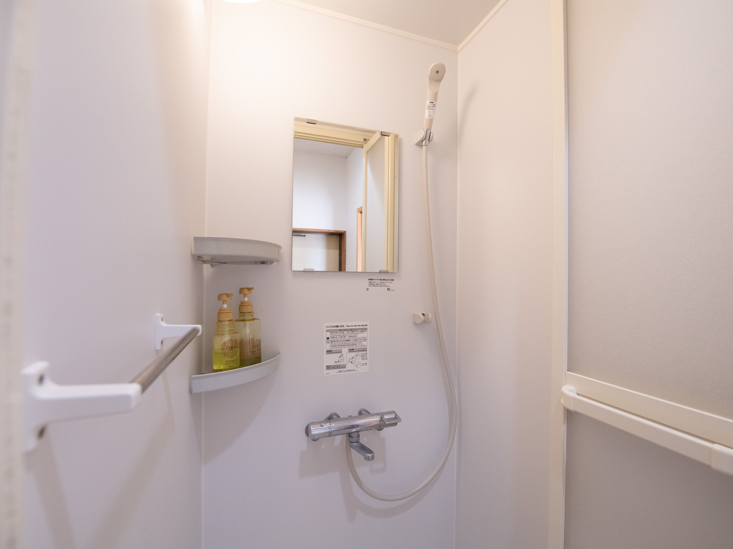 Une petite salle de bain blanche, équipée d’une douche, d’un miroir et d’une étagère où sont rangés les articles de toilette.