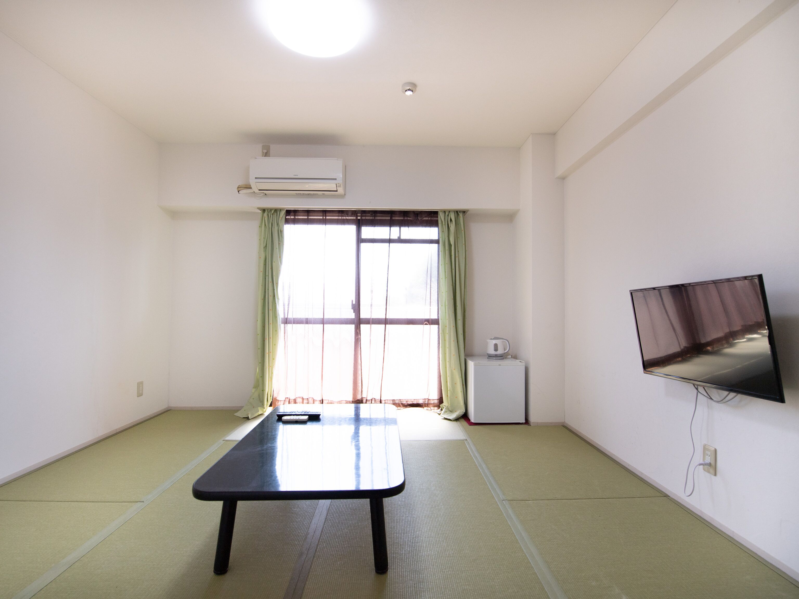A room with tatami flooring, a low table, a television mounted on the wall, and a window with curtains.