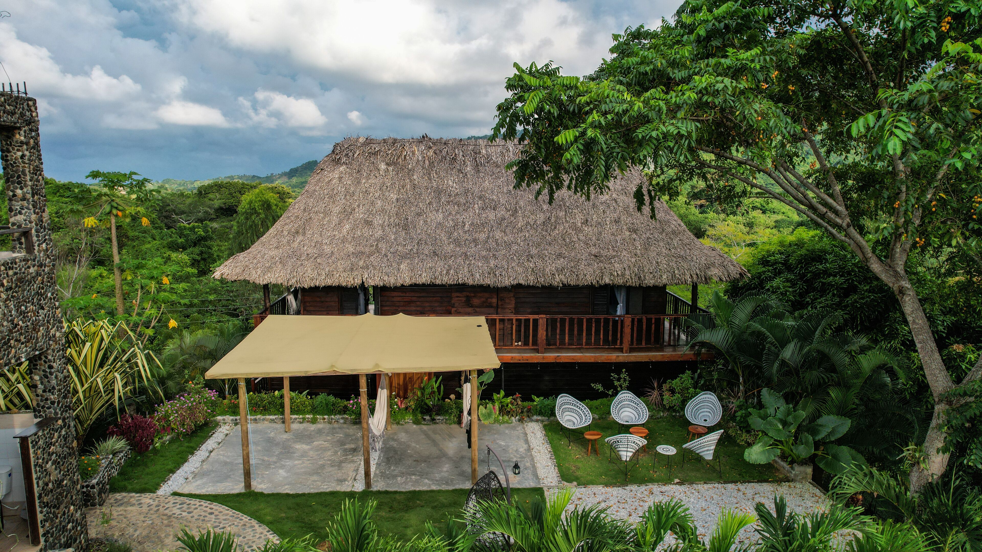 Une maison à toit de chaume dotée d’une terrasse en bois, entourée d’une végétation luxuriante et d’un jardin aménagé avec des espaces pour s’asseoir à l’extérieur.
