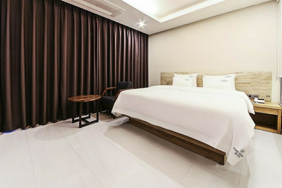 A modern hotel room with a large bed, dark curtains, a small round table, and a nightstand with a lamp.
