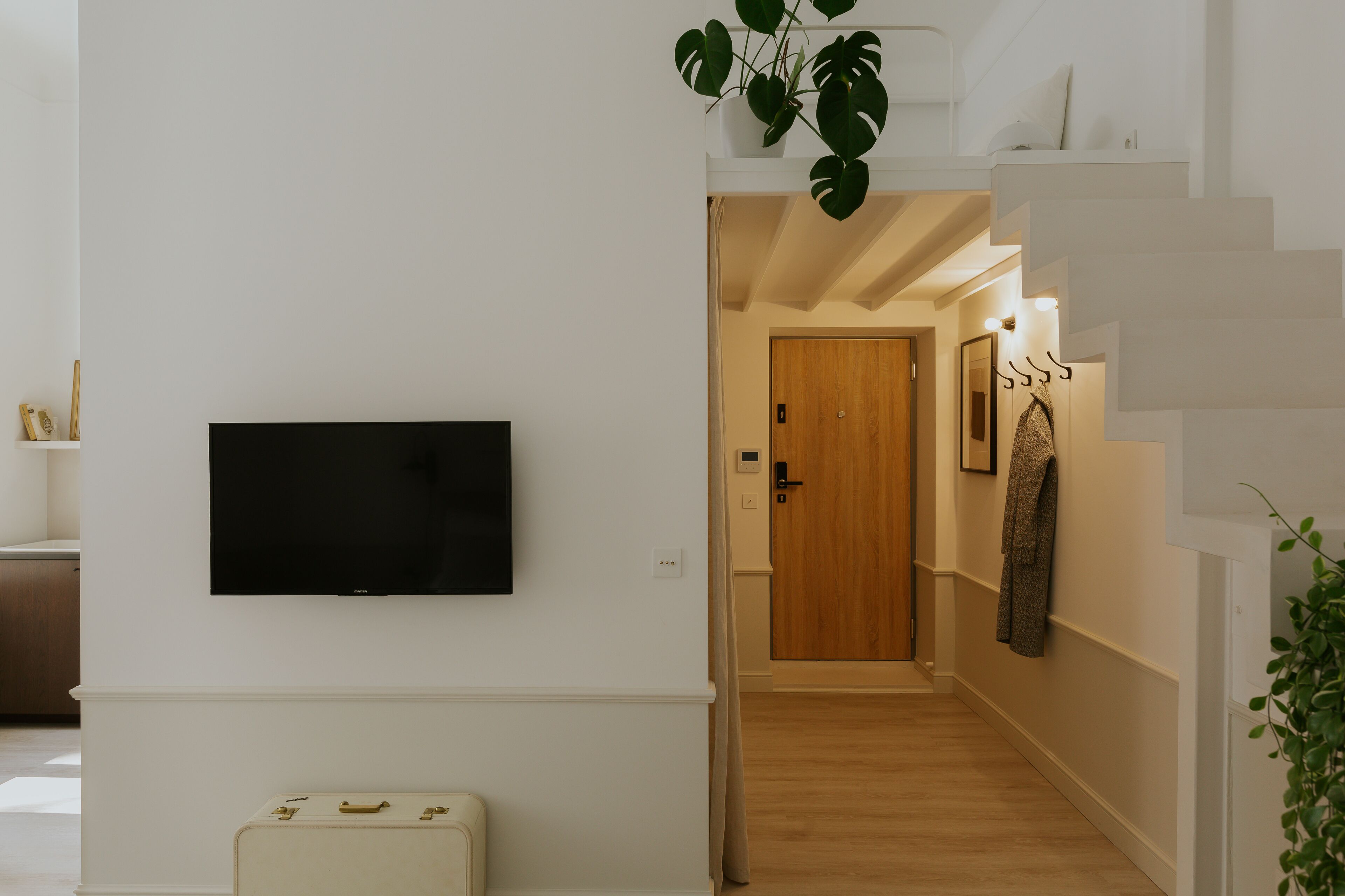 A hallway with a flat-screen TV mounted on the wall, a wooden door, and a coat rack with a jacket.