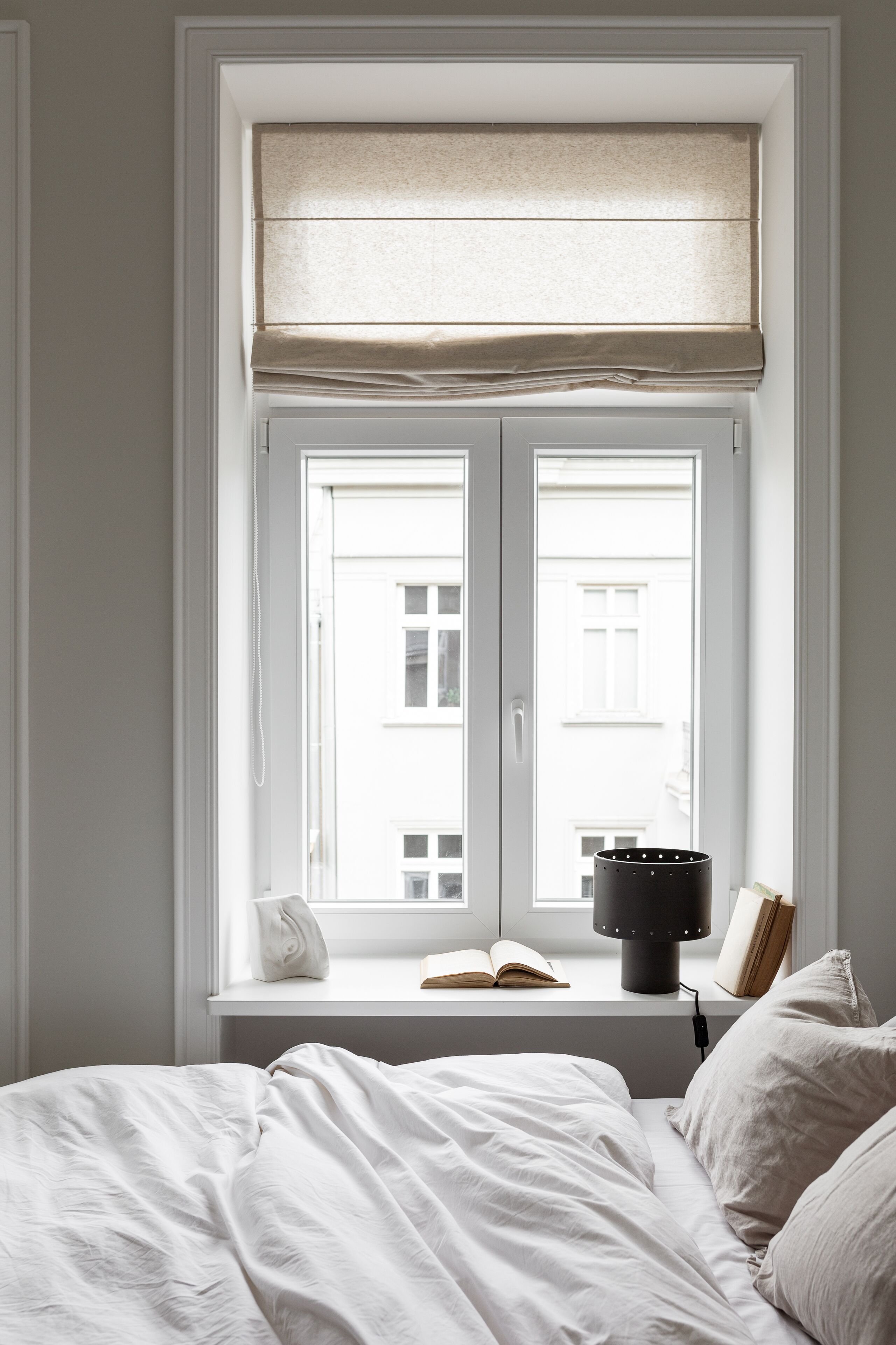 A bedroom with a large window, a bedside table with a lamp, and a book.