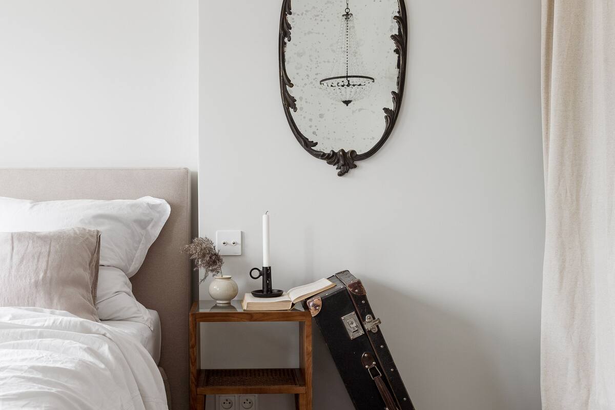 A bedroom with a bed, a bedside table with a book, a candle, and a suitcase leaning against the wall.