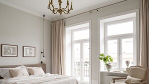 A bedroom with a large bed, a chair, a window with curtains, and a plant on a windowsill. - Lwowska Studios - STAYINWARSAW (Warsaw)