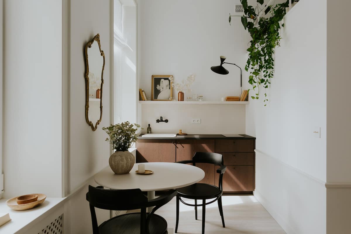 A dining area with a round table, two chairs, a vase with flowers, and a mirror on the wall.