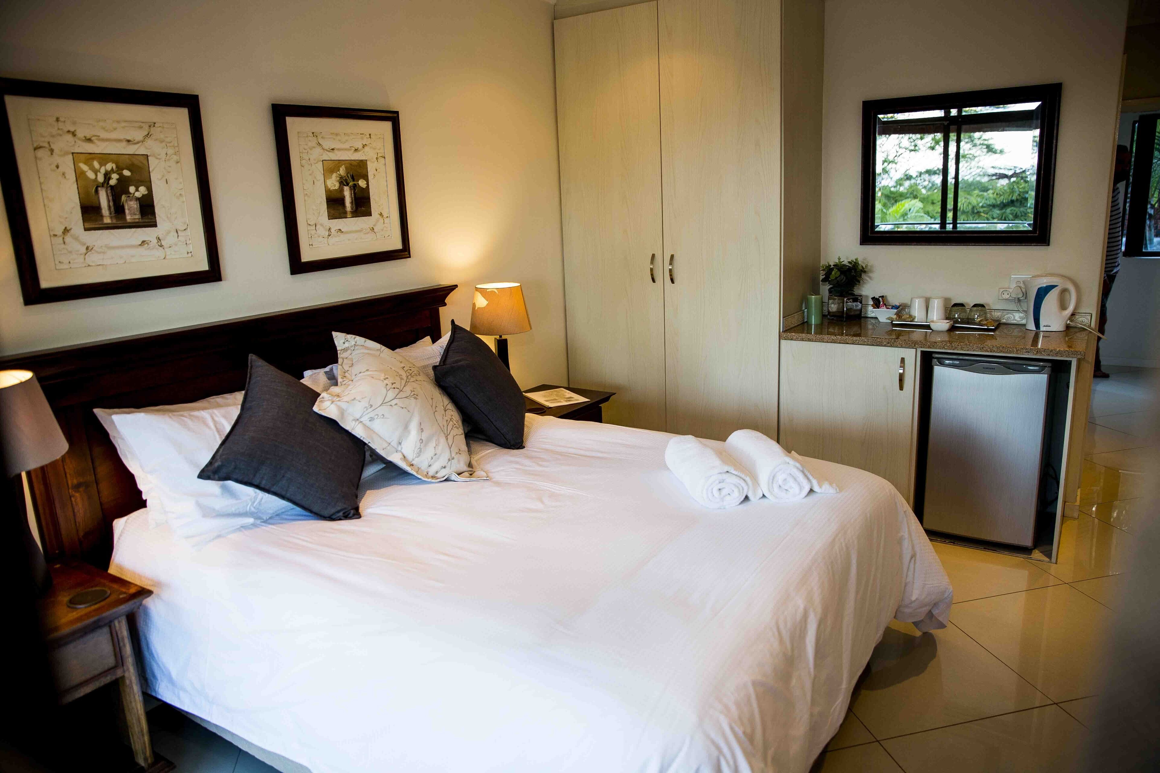 Una cama bien hecha con almohadas, una lámpara de noche, una mesita, un armario y una zona de kitchenette con ventana.