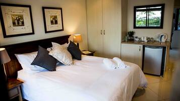 Una cama bien hecha con almohadas, una lámpara de noche, una mesita, un armario y una zona de kitchenette con ventana.