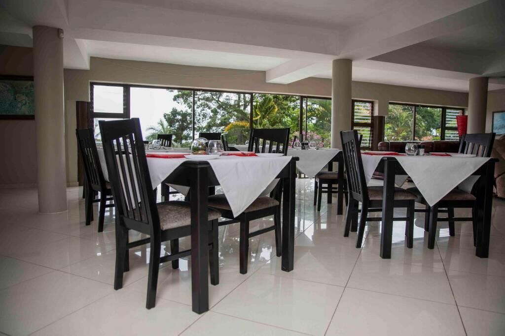 Uma área de refeições com mesas para quatro pessoas, rodeada por cadeiras, grandes janelas e vista para o verde.