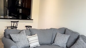 A gray sectional sofa with patterned pillows, a black coffee table with ornate legs, and a kitchen area in the background.