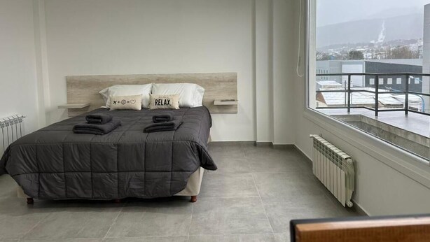 A modern bedroom with a large bed, a view of the city, and a window with a view of a building. - Jainen Apartamentos (Ushuaia)