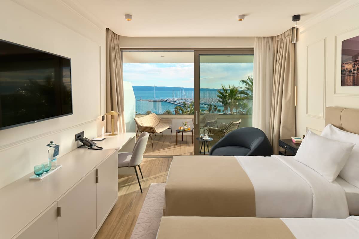 A hotel room with a bed, a desk, a chair, a TV, and a balcony with a view of the sea.