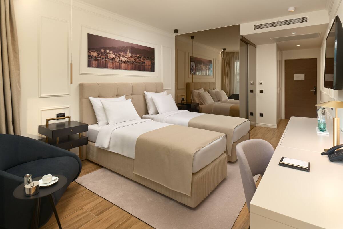 A hotel room with two beds, a desk, a chair, a small table with a cup and a pitcher, and a large mirror.
