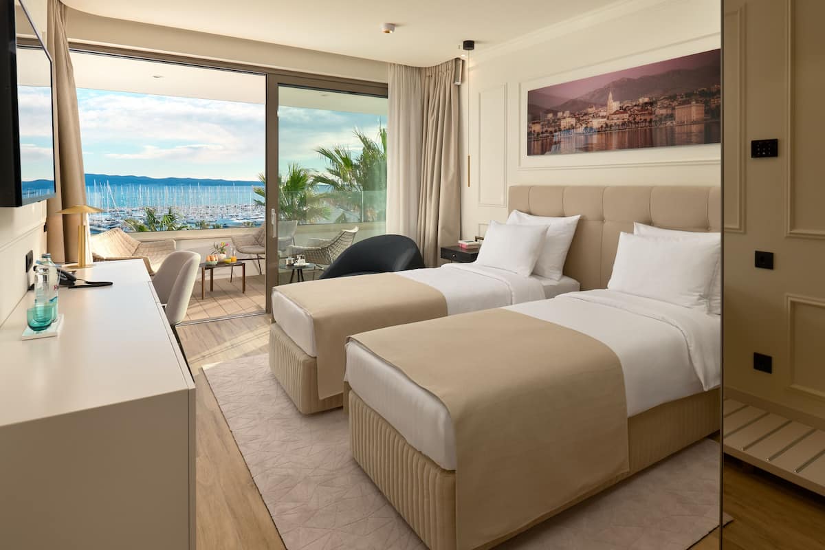 A hotel room with two beds, a desk, a chair, and a large window with a view of the sea.