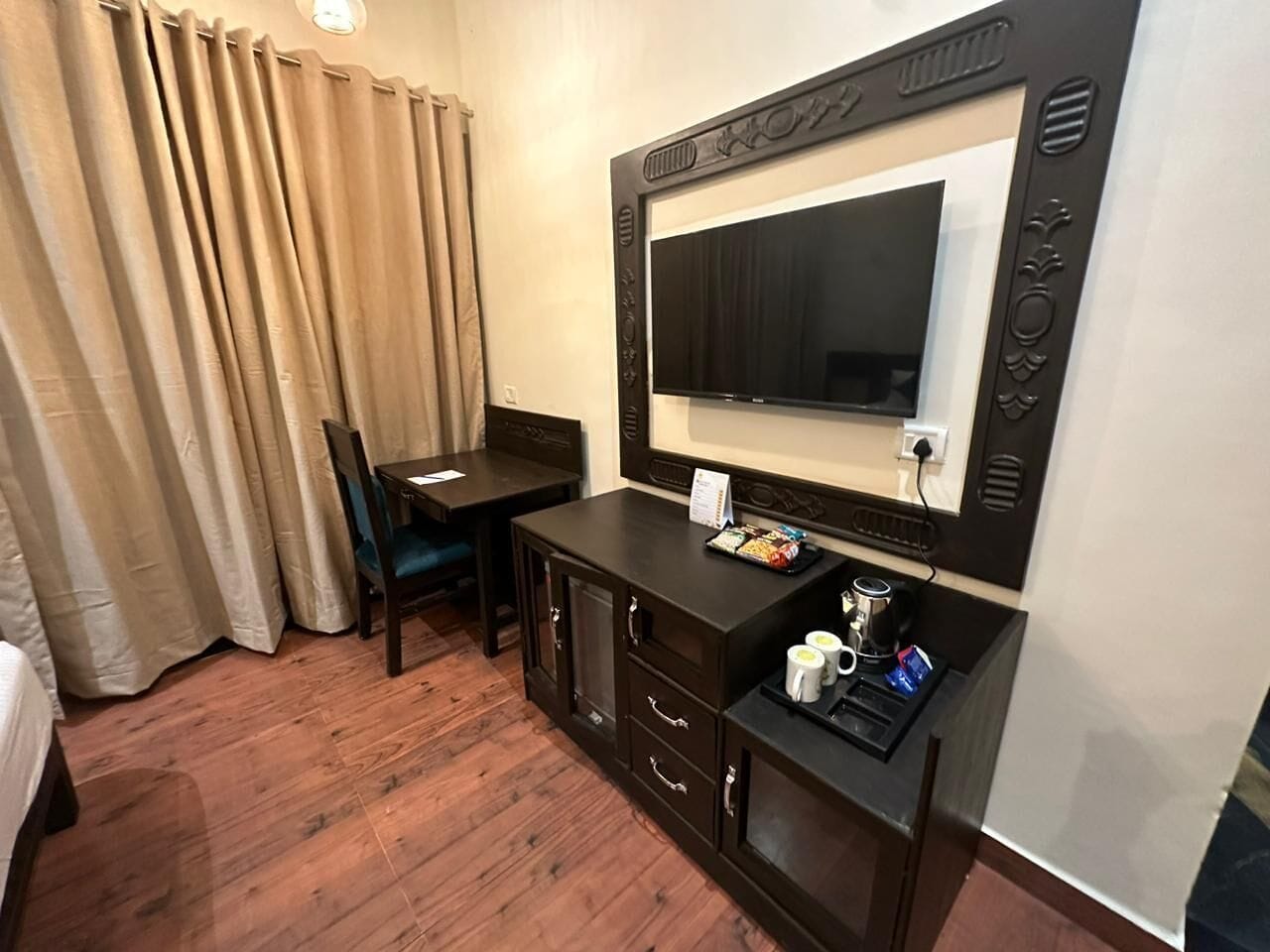 A hotel room with a wooden floor, a desk with a chair, a television, and a minibar.
