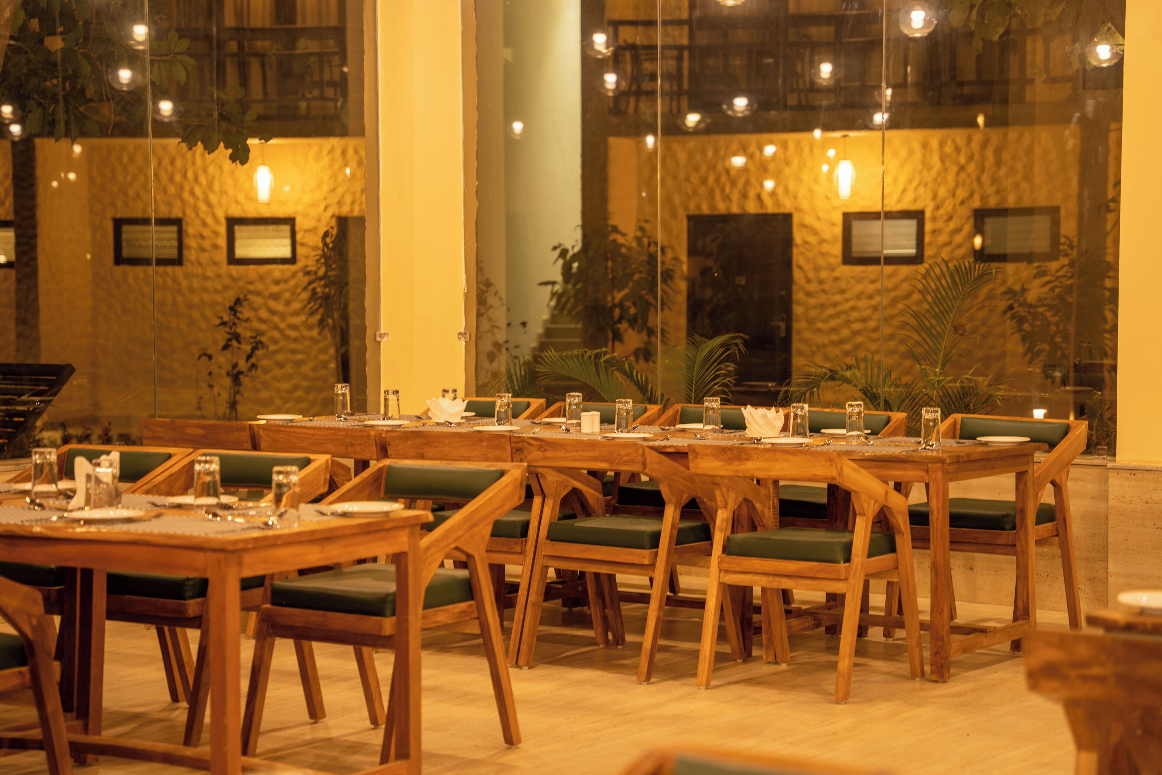 Un restaurante con mesas de madera y sillas con cojines verdes, dispuesto en un amplio comedor con vista al interior a través de un muro de cristal.