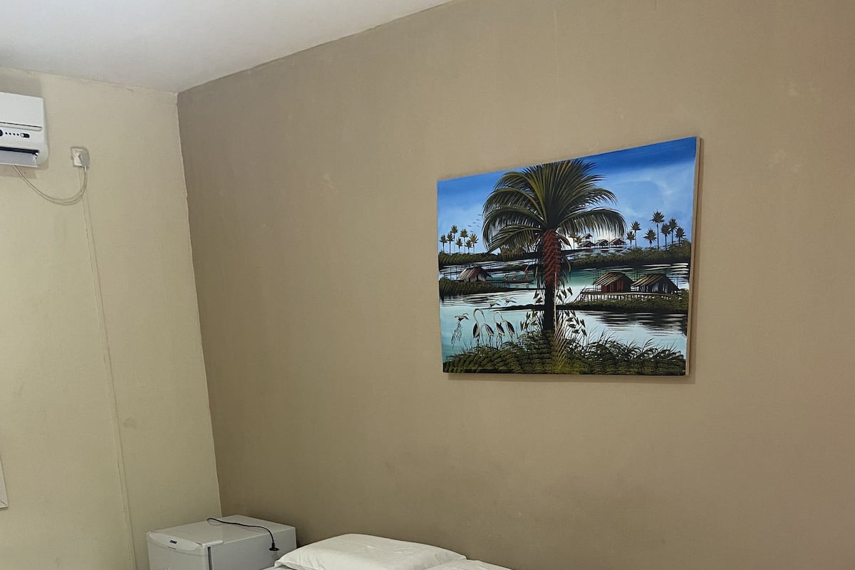 Habitació d'hotel amb dos llits, un quadre d'un paisatge tropical, un aparell d'aire condicionat blanc i una taula petita amb un comandament a distància.