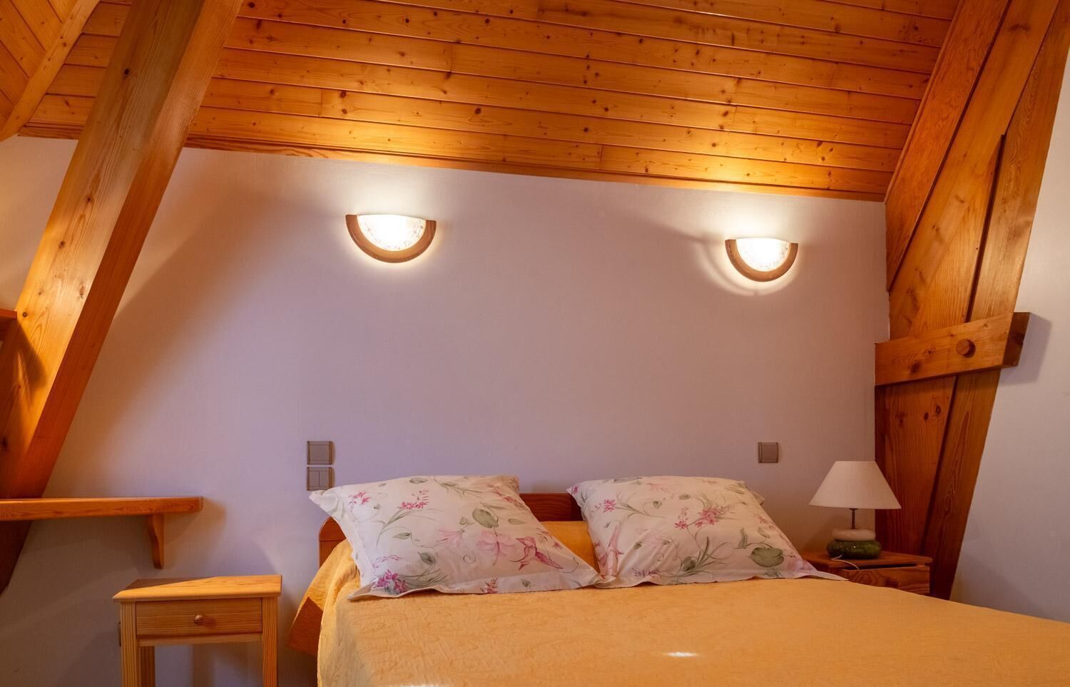 Une chambre avec un plafond en pente, des poutres en bois, deux lampes fixées au mur, un lit avec une literie à motifs floraux, une table de chevet avec une lampe et une petite table en bois.