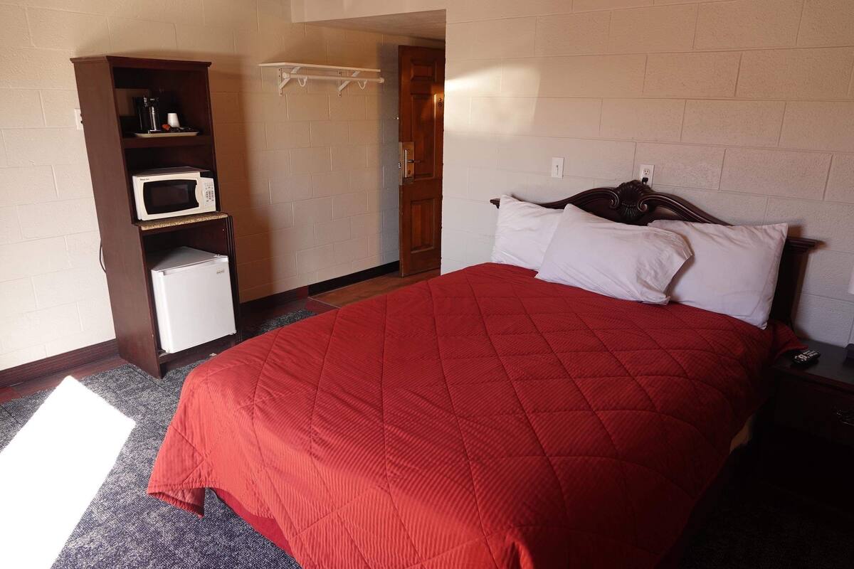 A bedroom with a red bedspread, a white pillow, a wooden headboard, a small white refrigerator, and a microwave on a shelf.