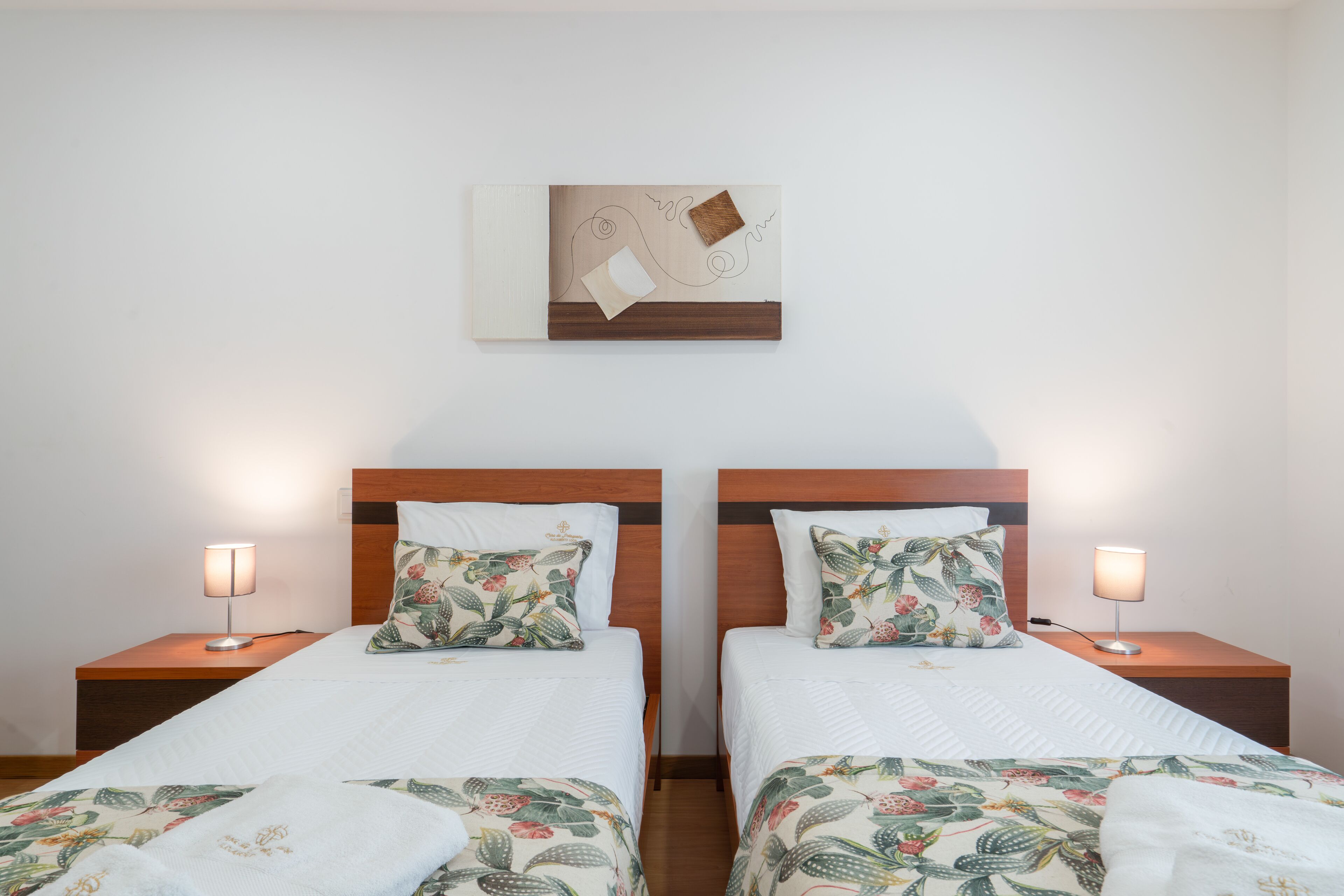 Quarto de hotel com duas camas, cada uma com colcha e almofadas estampadas com motivos florais. Possui cabeceira de madeira, uma obra de arte na parede e luminárias de cabeceira.