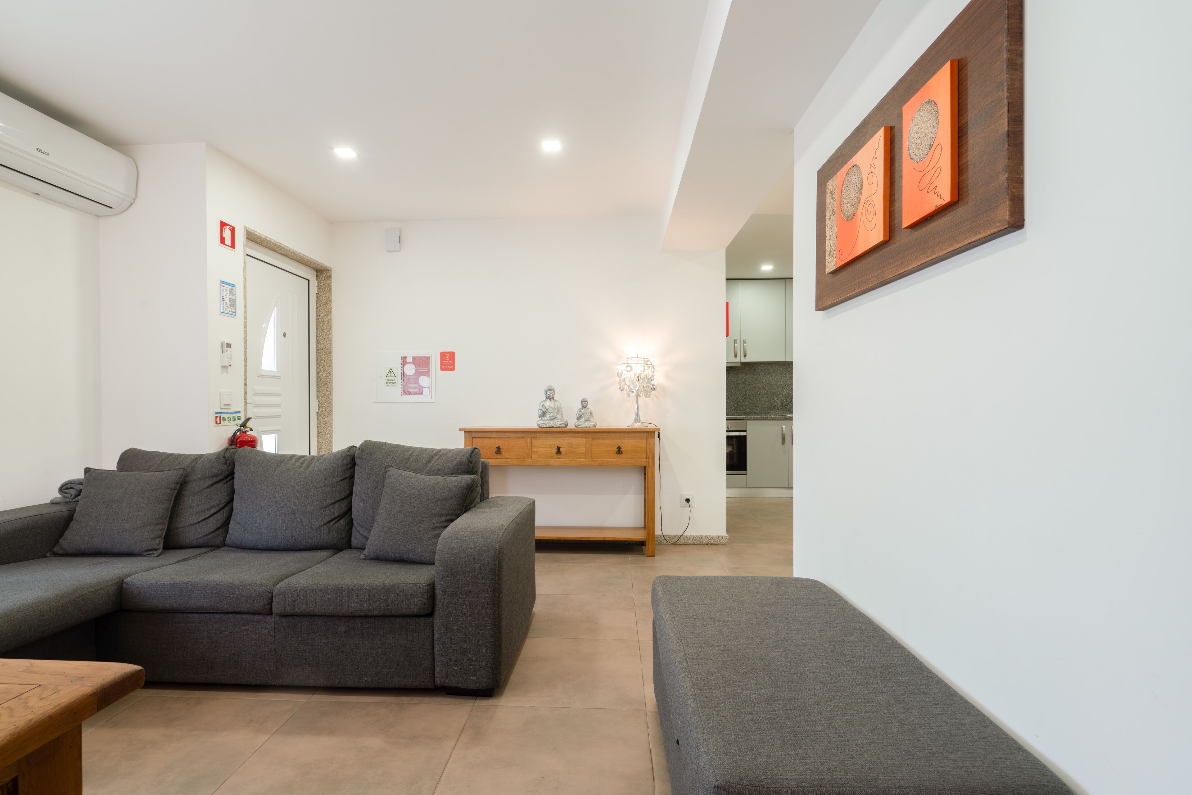 A modern living room with a grey sofa, a wooden coffee table, and a small side table. There is a wooden cabinet against the wall and a door with a fire alarm.