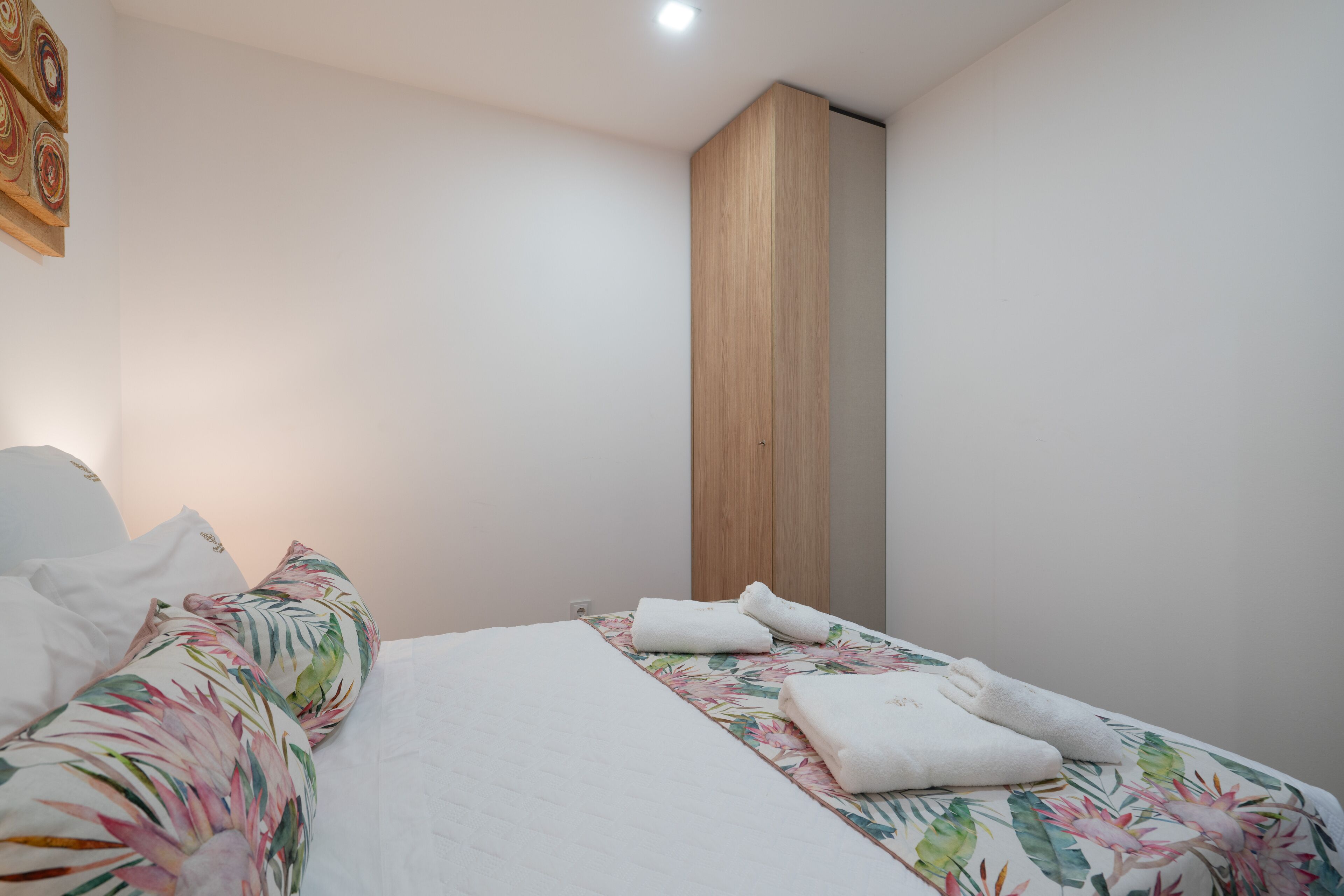 Un lit bien fait, avec une literie à motifs floraux et des serviettes blanches, une armoire en bois et un mur sobre agrémenté d’un seul luminaire.