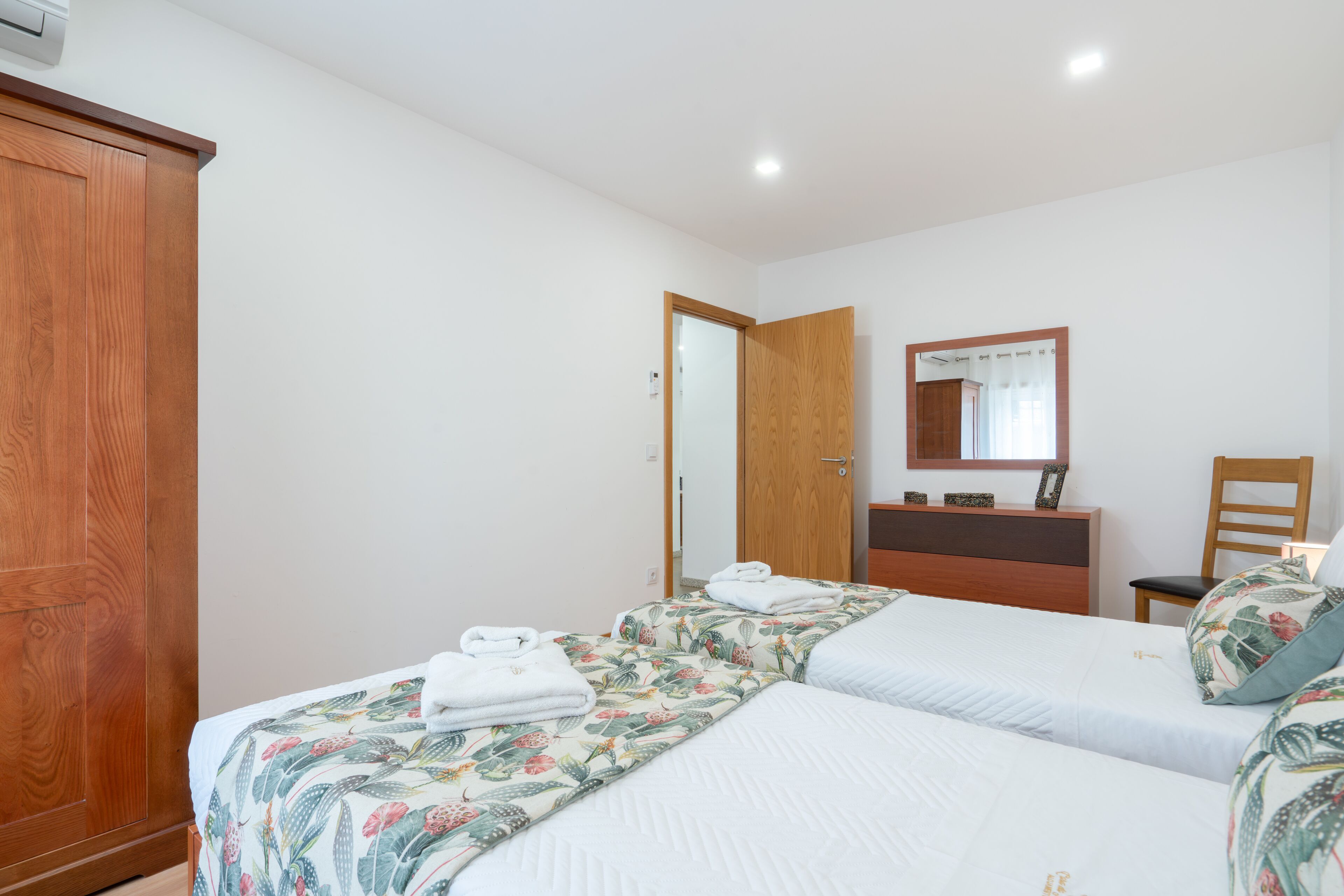 A hotel room with two beds, a wooden wardrobe, a mirror, and a desk with a chair.