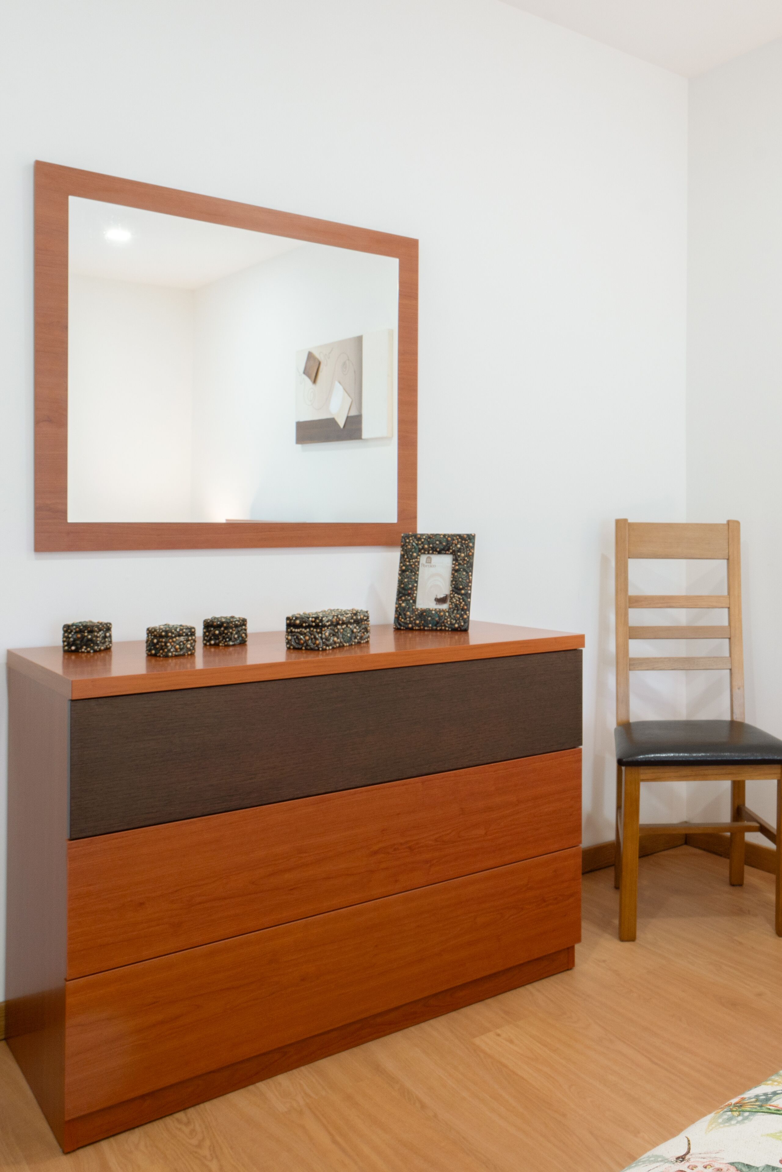Un aparador de madera con un espejo encima, una silla a la derecha y un cuadro en el aparador.