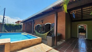 Ein Swimmingpool mit Hängematte und Gartenmöbeln, ein Ziegelgebäude mit überdachter Terrasse und ein gefliester Weg.