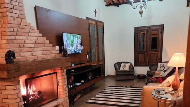 A cozy living room with a brick fireplace, a television mounted above it, and a comfortable seating area with a sofa and armchairs. - Magnífico Rancho em Jarinu (Jarinu)
