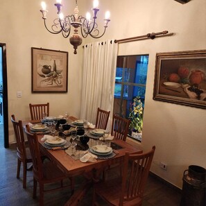 Uma sala de jantar com uma mesa de madeira posta para seis pessoas, rodeada por cadeiras. Há um candelabro sobre a mesa, um quadro emoldurado na parede e uma janela com cortinas.