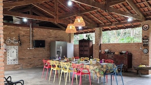 A spacious dining area with a high ceiling and exposed beams, featuring a rustic brick wall, a large wooden cabinet, and colorful chairs around a table set for dining. - Magnífico Rancho em Jarinu (Jarinu)