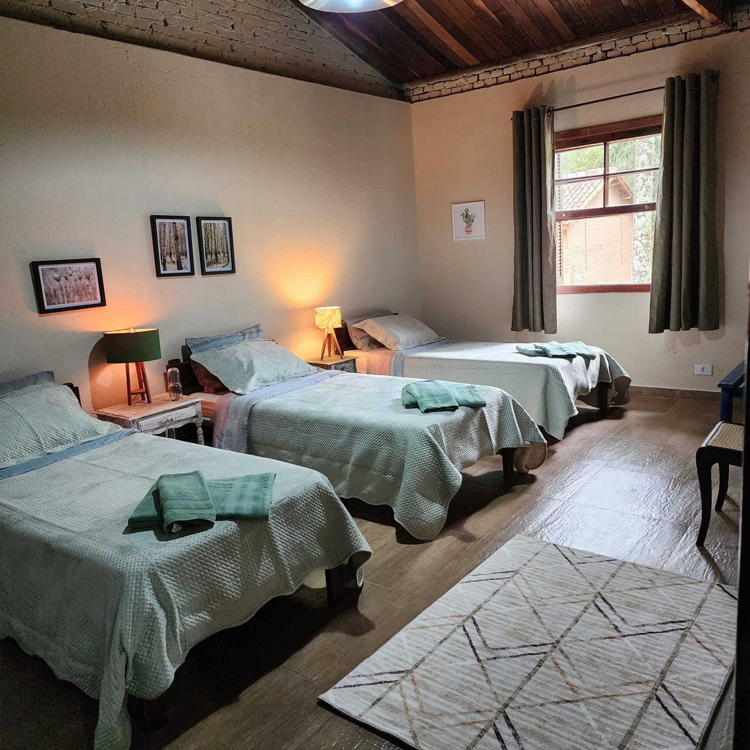 A room with three beds, each with a green blanket, a small table with a lamp, and a window with curtains.