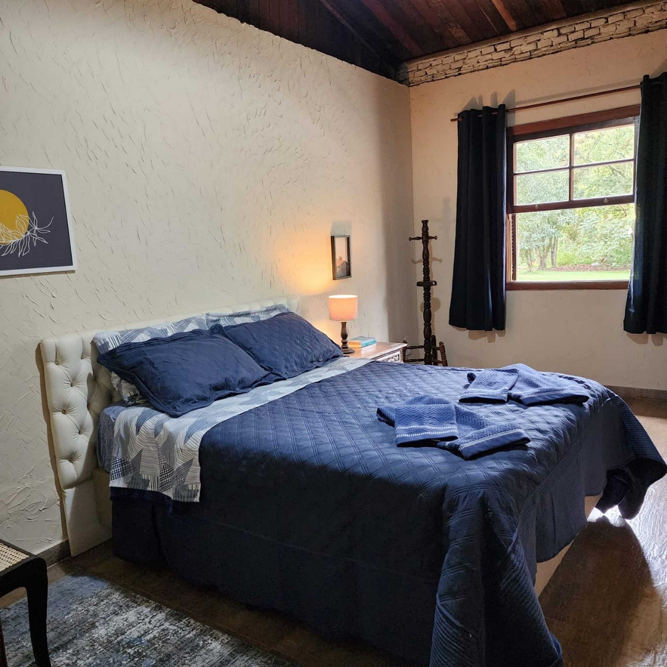 Un dormitorio con cama, mesita de noche, ventana con cortinas e imagen colgada en la pared.