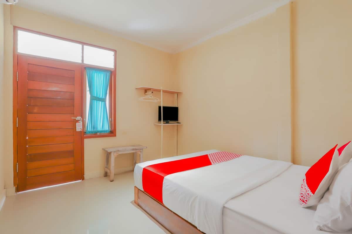 A bedroom with a bed, a wooden door, a small table, a TV on a stand, and a window with curtains.