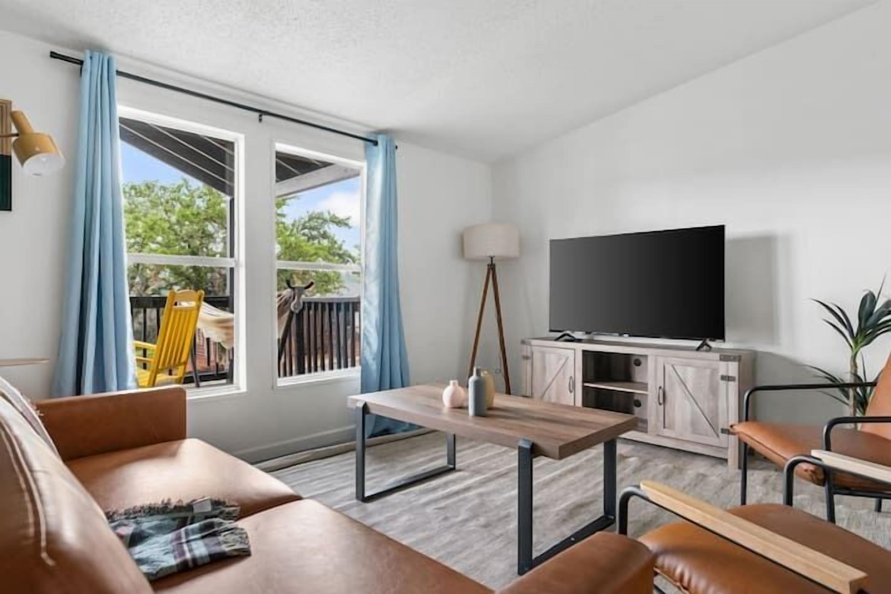 A modern living room with a sofa, coffee table, and TV. There is a window with curtains and a view of greenery outside.