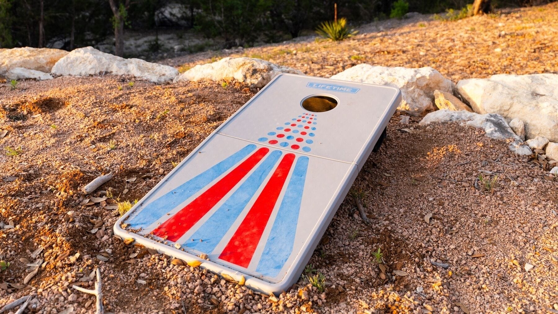 Een jeu de boules baan met een rood-blauw streepdessin, op een grindoppervlak met stenen en schaarse begroeiing eromheen.