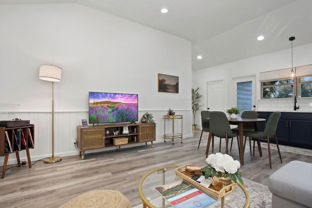 A modern living room with a flat-screen TV, a floor lamp, a dining area with chairs, and a kitchen with cabinets.