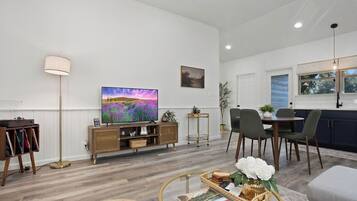 A modern living room with a flat-screen TV, a floor lamp, a dining area with chairs, and a kitchen with cabinets.