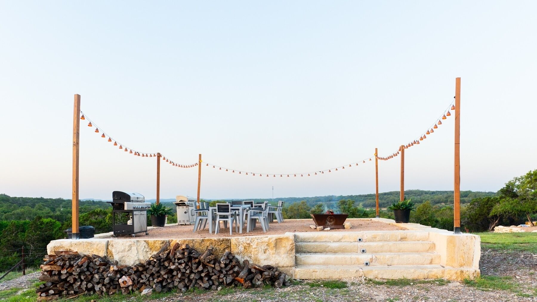 An outdoor seating area with a grill, chairs, and a fire pit, surrounded by a stone platform and a view of the countryside.