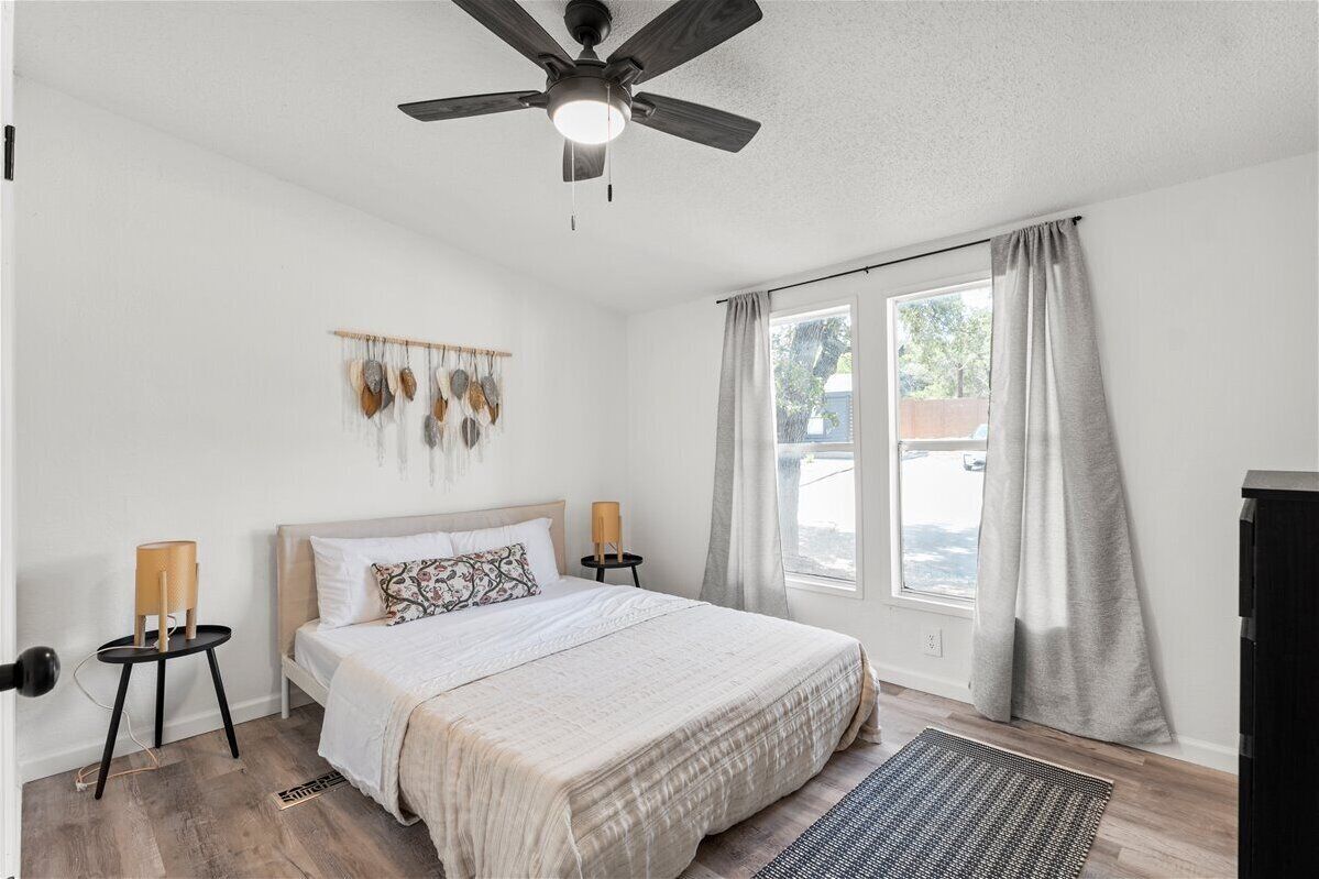 Une chambre à coucher comprenant un lit, un ventilateur de plafond, une fenêtre avec des rideaux, un luminaire fixé au mur et une petite table avec une lampe.