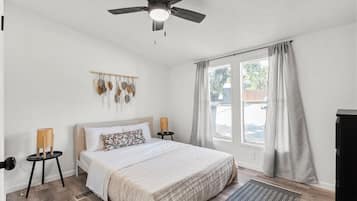 Une chambre à coucher comprenant un lit, un ventilateur de plafond, une fenêtre avec des rideaux, un luminaire fixé au mur et une petite table avec une lampe.