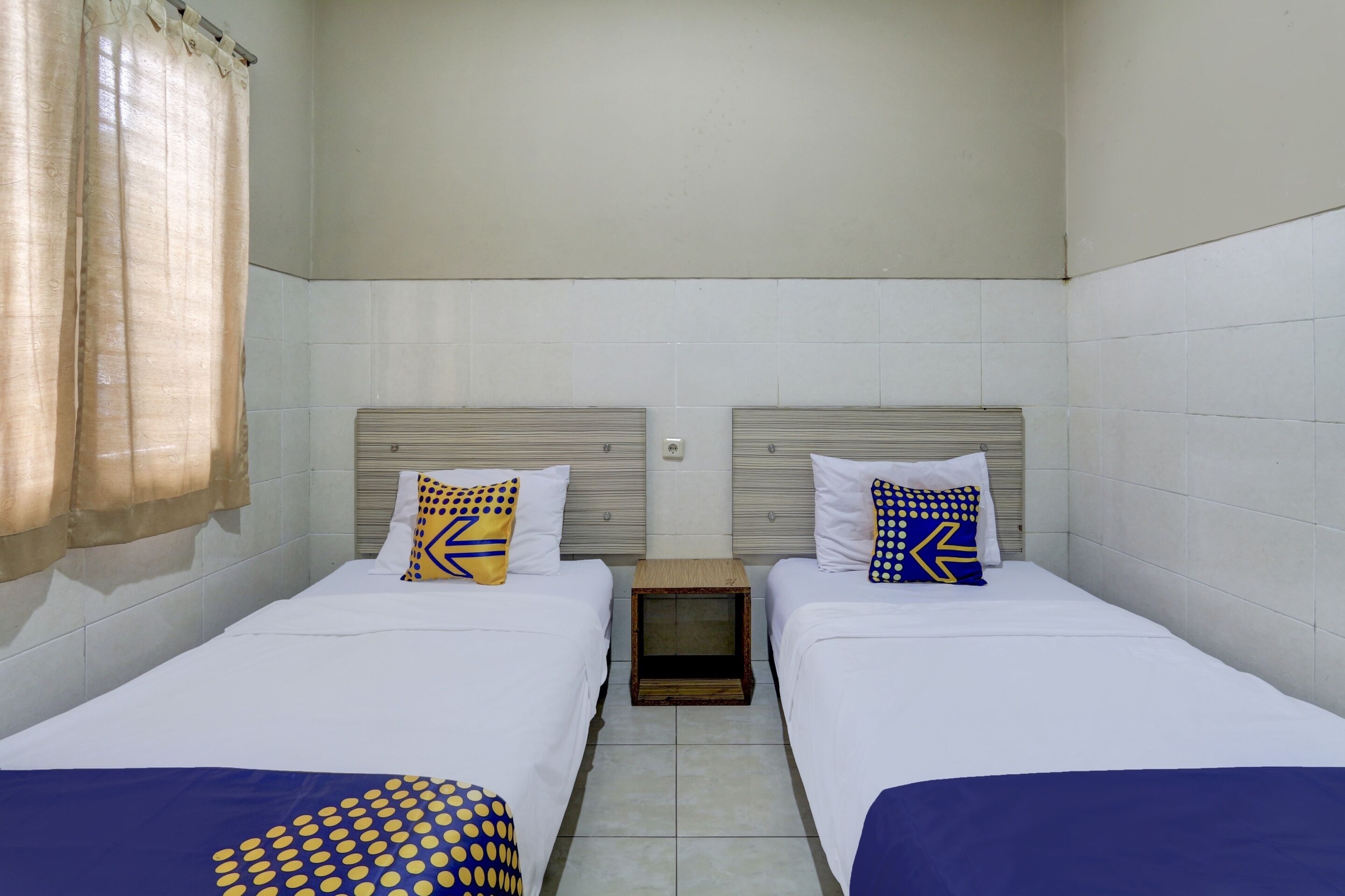 A small, clean room with two beds, each with a pillow and a blanket. There is a small wooden nightstand between the beds.