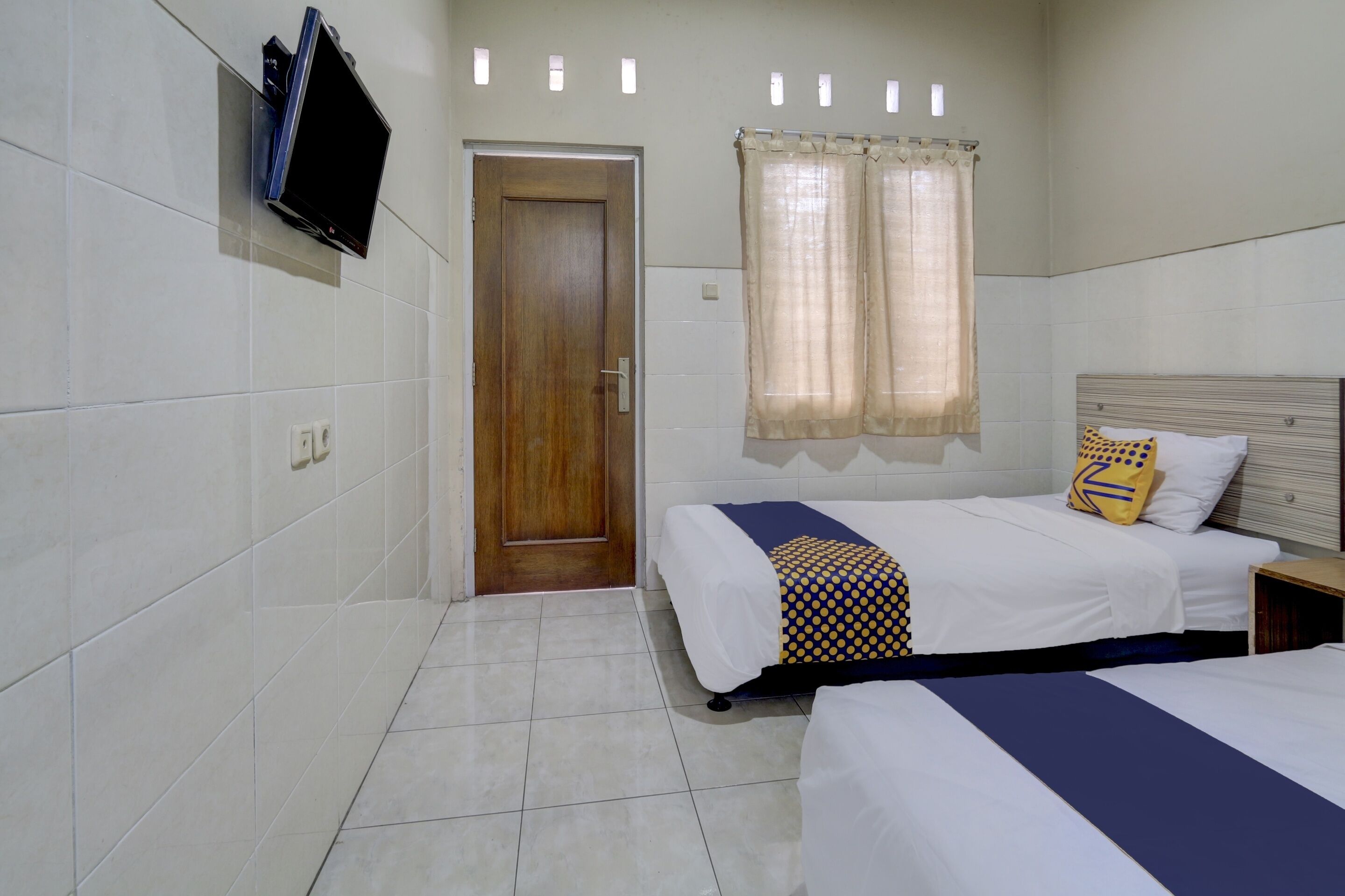 Habitación de hotel con dos camas, una puerta de madera, un televisor montado en la pared y una ventana con cortinas.