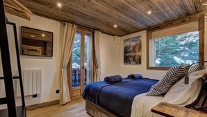 Ein Schlafzimmer mit Holzboden, einem großen Fenster mit Blick auf einen Wald, einem Bett mit dunkler Bettwäsche und einem Heizkörper an der Wand.