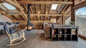 Ein gemütlicher Loftsraum mit hohen Decken, freiliegenden Holzbalken, Kamin, Schaukelstuhl, einer Couch und einem Bücherregal.